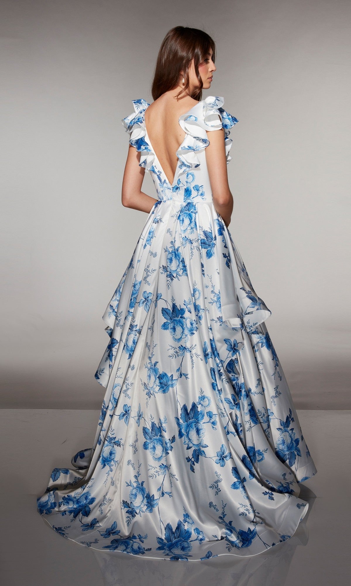 A woman stands backwards against a neutral background wearing the Alyce 62001 long prom dress, featuring satin fabric, blue floral print, ruffled sleeves, and a deep V-back.